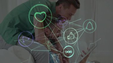 Erkek çift, yüzen sosyal medya simgelerinin altını çizerek ev teknolojisi sahnesinde tablet ekranına yaslanıyor. Duygu, yakınlık, bağlantı, dijital iletişim, modern yaşam tarzı, kutlama, rahatlama