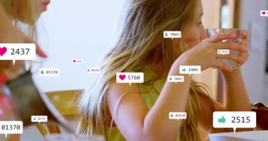 Kahvaltıda elinde kaşık tutan ve bardakları kaldıran iki kız yüzen dijital pazarlama ikonları gösteriyor. Çocuklar, kahvaltı, sosyal medya, tepkiler, dijital etkileşim, aile, oyun