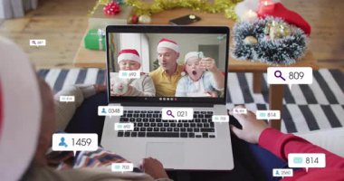 Kırmızı kazaklı ve Noel Baba şapkalı biri kanepede dizüstü bilgisayar kullanıyor, sosyal medya simgelerini paylaşıyor. Şenlik, tatil, aile, kutlama, bağlantı, sıcak, sıcaklık