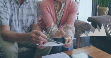 Üst düzey bir çift ev lobisinde finans okumak için öne doğru eğiliyor ve animasyon bar çizelgeleriyle örtüşüyor. Emeklilik, mali planlama, yaşlılık, teknoloji, ev ortamı, analiz, konfor