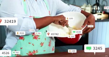 Üst düzey kadın mutfak masasında karışımı karıştırıyor, pazarlamayı yüzen bildirim simgeleriyle gösteriyor. Yaşlılar, ev hanımları, canlı, dijital, yerli, teknoloji, gelenek