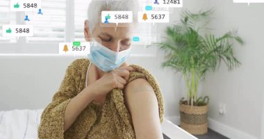 Kıdemli maskeli kadın kollarını kaldırıyor. Aşı bandajını gösteriyor. Yüzen sosyal simgeler oluşturuyor. Yaşlılar, sağlık, sağlık, pozitiflik, sosyal medya, toplum, koruma