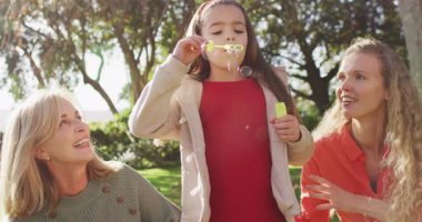 Video of happy caucasian daughter blowing bubbles with mother and grandmother in autumn garden. Family, domestic life and togetherness concept digitally generated video.
