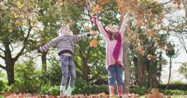 Video of happy caucasian brother and sister having fun throwing autumn leaves in garden. Family, domestic life and togetherness concept digitally generated video.