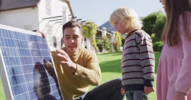 Video of happy caucasian father explaining solar panel to son and daughter in sunny garden. Family, domestic life and togetherness concept digitally generated video.