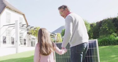 Video of happy caucasian grandfather carrying solar panel walking with granddaughter in sunny garden. Family, domestic life and togetherness concept digitally generated video.