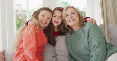 Video portrait of happy caucasian mother, grandmother and granddaughter embracing on couch. Family, domestic life and togetherness concept digitally generated video.