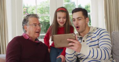 Video of happy caucasian father, grandfather and granddaughter sitting on couch looking at tablet. Family, domestic life and togetherness concept digitally generated video.