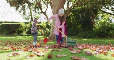 Video of happy caucasian brother and sister raking up autumn leaves in sunny garden. Family, domestic life and togetherness concept digitally generated video.