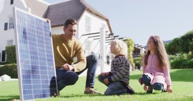 Video of happy caucasian father explaining solar panel to son and daughter in sunny garden. Family, domestic life and togetherness concept digitally generated video.