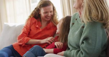 Video of happy caucasian daughter tickled by mother and grandmother sitting on couch. Family, domestic life and togetherness concept digitally generated video.