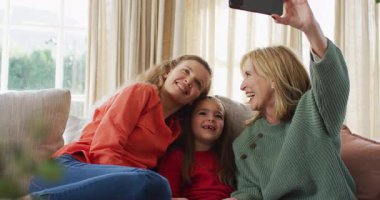 Video of happy caucasian mother and grandmother looking at smartphone with granddaughter on couch. Family, domestic life and togetherness concept digitally generated video.