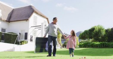 Video of happy caucasian grandfather carrying solar panel walking with granddaughter in sunny garden. Family, domestic life and togetherness concept digitally generated video.