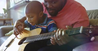Baba çocuğun elini gitar boynuna koyuyor ve gökkuşağının altında müzik dersi veriyor. Aile, akıl hocalığı, müzik, uyum, yetiştirme, sıcaklık, iç mekan.