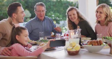 Video of happy caucasian parents, daughter and grandparents at dinner table looking at smartphone. Family, domestic life and togetherness concept digitally generated video.