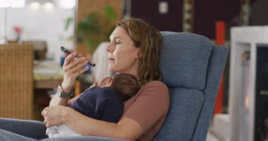 Kafkasyalı annenin yeni doğmuş bir bebekle koltukta oturup akıllı telefon kullandığı bir video. annelik, ebeveyn sevgisi ve yeni doğan bebek konseptiyle ilgilenmek dijital olarak üretilmiş video.