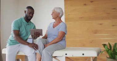 Afrika kökenli Amerikalı erkek fizyoterapistin beyaz yaşlı bir kadınla tablet kullanırken videosu. Yaşlılar sağlığı ve huzurevi konsepti dijital olarak oluşturulmuş video.