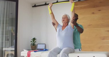 Afrika kökenli Amerikalı erkek fizyoterapistle egzersiz yapan mutlu, beyaz bir kadının videosu. Yaşlılar sağlığı ve huzurevi konsepti dijital olarak oluşturulmuş video.