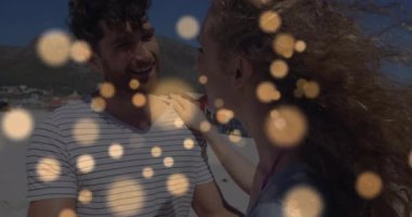 Bir kadın, erkeğin omzuna elini koyuyor, kumsaldaki kulübelerin yanında altın bokeh ile dans ediyor. Romantizm, dans, deniz kenarı, eğlence, coşku, sükunet, bağlantı