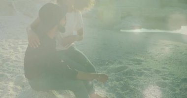 Güneş ışığı parlayan grafikleri tetiklerken diz çökmüş çiftin kıyı şeridinde kucaklaşmalarını gösteren bir video. Romantizm, samimiyet, bağlantı, doğa, günbatımı, huzur, şefkat