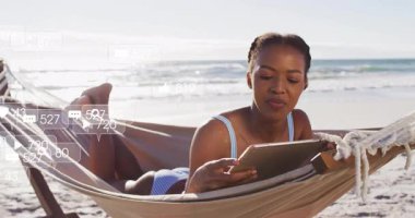 Deniz kenarındaki hamakta tablet kullanan bir kadın kulaklıkla dinleyen baloncuklarla sosyal istatistikleri kontrol ediyor. Rahatlama, dijital, açık hava, eğlence, yaşam tarzı, teknoloji, dinginlik