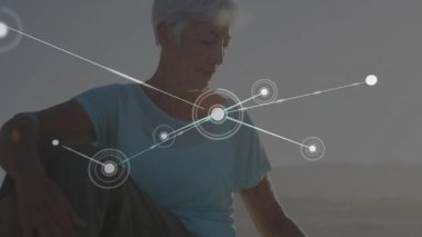 Yaşlı kadın aşağıya bakıyor, parlayan düğümleri etkinleştiriyor ve plajdaki sağlık ağını haritalıyor. Gerontoloji, farkındalık, bağlantı, dinginlik, dijital arayüz, refah, rahatlama
