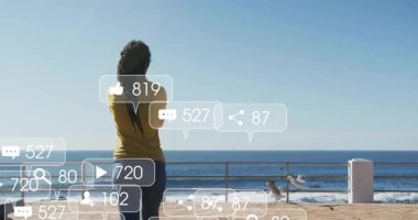 Beachfront woman gazing ocean receiving growing social media icons and raising arms for engagement. Voyage, solitude, contemplation, connection, digital, tranquility, nature