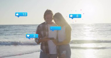 Günbatımında kumlu sahilde akıllı telefonları dinleyen bir çift sosyal medya simgeleri oluşturuyor. Romantizm, teknoloji, iletişim, eğlence, açık hava, canlı, bağlantı