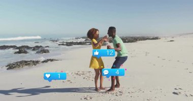 Kumsalda el ele tutuşan bir çift sosyal medya simgeleri ve konfeti güncellemesi yapıyor. Romantizm, kutlama, dijital, sevgi, yolculuk, canlı, açık hava