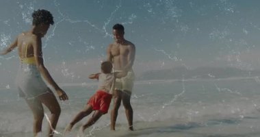 Baba, çocuğu elleriyle kaldırıyor ve annesi seyahat reklamına katılırken sığ dalgalarda sallanıyor. Aile, sahil, tatil, eğlence, neşe, açık hava, kaynaşma