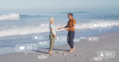 Sosyal medya simgeleri yükselirken kıdemli çift kumsalda el ele dans ederek kucaklaşıyor. Romantizm, bağlantı, uyum, şefkat, sükunet, boş zaman, açık hava