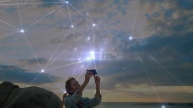 Sahilde diz çökmüş akıllı telefonu gökyüzüne doğru kaldıran dijital ağı aktifleştiren, gün batımını izleyen bir kadın. Doğa, teknoloji, bağlantı, sükunet, açık hava, yenilik, keşif