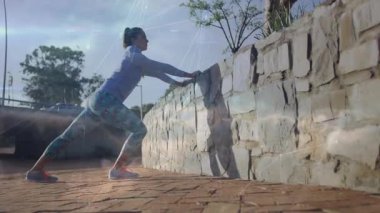 Güneşli avluya giren kadın ellerini taştan duvara koyuyor dizlerini bükerek spor yapmak için ittiriyor. Fitness, egzersiz, aktif giyim, kentsel, açık hava, dinamik, motivasyon