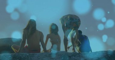 Şortlu şortlar, mavi Bokeh sürüklenirken arkadaşlarının yanına çömelip deniz manzarası seyrediyor. Macera, yoldaşlık, eğlence, doğa, sükunet, manzara, açık hava.