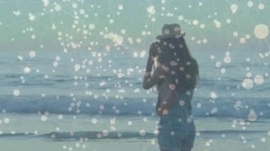 Fötr şapka takan bir kadın sahil şeridinde kamera kaldırıyor, ufku çerçeveliyor ve seyahat görüntülerini yakalıyor. Kıyı şeridi, keşif, dinginlik, rahatlık, açık hava, manzara, macera