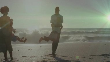 Aile, gün batımının parladığı bir ortamda kumlu sahilde çocukları döndürmek için tek ayak üzerinde dönüyor. Aileler, açık hava, eğlence, uyum, hareket, neşe, bağlantı
