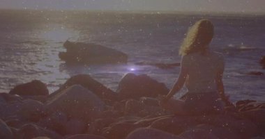 Sağlığı için kayalık kıyı şeridinde meditasyon yapan bir kadın. Güneş ufukta belirdiğinde merceklerin parlamasına yol açıyor. Dinginlik, farkındalık, doğa, manzara, sağlık, sükunet, açık hava