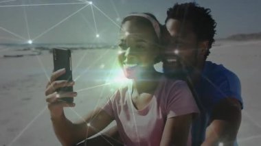 Kadın telefonu kaldırıyor ve kumsalda poz veren çift titreşen düğümlerle selfie çekiyor. Romantizm, bağlantı, teknoloji, plaj, mutluluk, açık hava, canlı