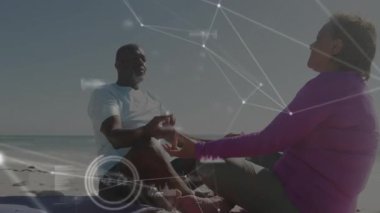 Olgun bir çift kumların üstünde el ele tutuşup meditasyon yapıyor, sağlık ölçümlerini gösteren animasyon ağlarını soluyor. Refah, rahatlama, bağlantı, farkındalık, dinginlik, dijital, uyum