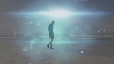 Adam kumun üzerine basıyor kürelerin iz sürmesine, teknoloji gösterilerinde kabuk büküyor. Yalnız figür, kıyı şeridi, soyut desenler, çevresel ışıltı, dijital ışık, düşünceli, minimalist