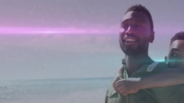 Baba, oğlunu kumsalda sırtında taşıyarak çerçeveye giriyor. Aileyi kutlayan mercekler üretiyor. Aile, bağ, açık hava, güneş ışığı, boş zaman, bağlantı, mutluluk.