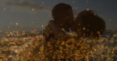 Çiftler okyanusta sarılıp öpüşürken altın parıltılar seyahat sahnesini ortaya çıkarmak için yanıp tutuşuyor. Romantizm, samimiyet, sükunet, siluet, kutlama, şefkat, sükunet