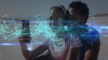 Kadın ve erkek kumsalda oturmuş telefon çekiyor selfie çekiyor dalga formlarını ateşliyor ve teknolojiyi ateşliyor. Romantik, açık hava, aydınlık, modern, bağlantı, rahatlama, uyum