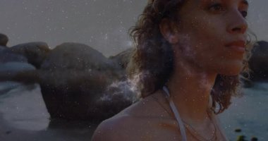 Sahilde duran bir kadın kulağının arkasında nebula yapıyor. Yüzünde alevler var. Kozmik, dinginlik, manzara, mistik, düşünceli, ruhani, manzaralı