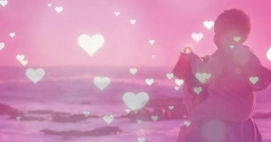 Su kenarına gelen bir kadın, okyanus dalgaları üzerinde atan kalplerle sağlıklı olmak için bebeği omuzlarına alıyor. Annelik, aile bağı, sahil, dinginlik, şefkat, şefkat, duygusal