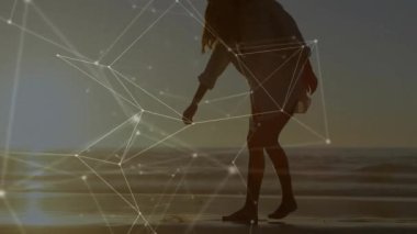 Kadın, dijital ağın altında kıyı şeridinde yürürken durup deniz kabuğu seçiyor ve köpek yanından geçiyor. Sahil, gün batımı, kıyı şeridi, soyut, teknoloji, yalnızlık, sükunet