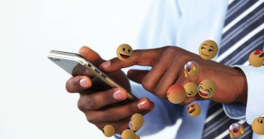 Stüdyoda açık mavi gömlekli ve kravatlı, yüzen emojilerle akıllı telefonları dinleyen adam. Teknoloji, iletişim, dijital, ifade, modern, iş, yenilik