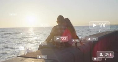 Çift, okyanus batımında deniz kenarında oturmuş nişan büyümesini gösteren sosyal simgeleri izliyorlar. Romantik, dinginlik, sükunet, dijital, bağlantı, ufuk, doğa