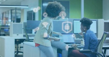 Dizüstü bilgisayarlardaki güvenlik simgelerini inceliyor, iki profesyonel modern açık planlı ofiste masanın üstüne yaslanıyor. İşbirliği, takım çalışması, teknoloji, modernlik, dijital güvenlik, yenilik, profesyonel