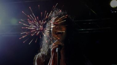 Kadın şarkıcı mikrofona konser sahnesinde vokal yapıyor ve arkasından havai fişek gösterisi yapıyor. Sanatçı, eğlence, dinamik, canlı, kutlama, sahne, gösteri